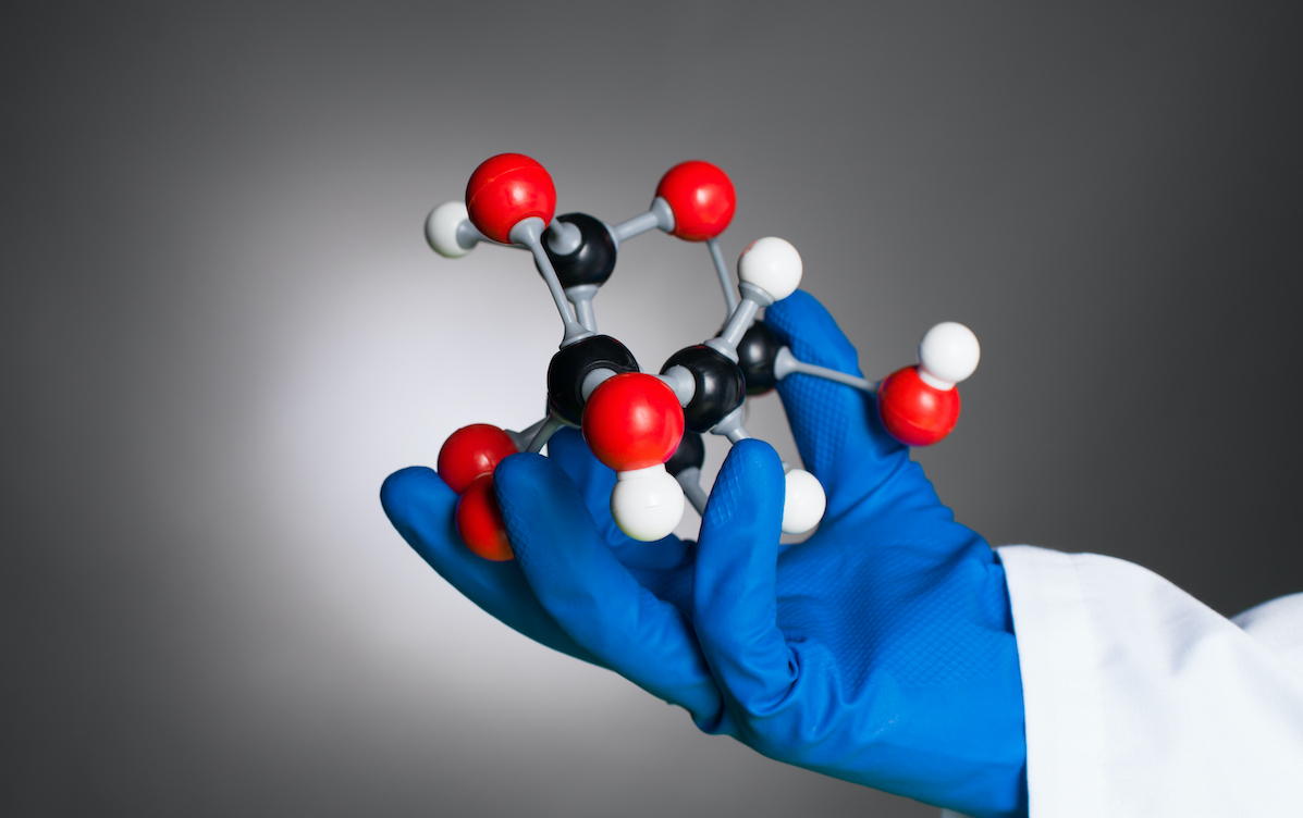 hand in blue rubber glove and white lab coat holding a 3d model of a mollecular structure made of white, black and red spheres with grey bonds, against a gradient grey background
